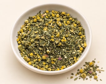 May include: A white bowl filled with a loose leaf herbal tea blend containing dried green leaves, small yellow chamomile flower heads, and tiny pink flower petals. A small pile of the same dried herbal tea mixture sits outside the bowl.