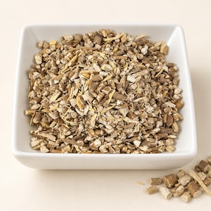 May include: A square white bowl filled with a mound of dried, chopped herbal root pieces. The pieces are a mix of light tan, beige, and dark brown woody textures. A small pile of the same dried root fragments sits outside the bowl to the lower right.