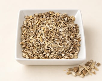 May include: A square white bowl filled with a mound of dried, chopped herbal root pieces. The pieces are a mix of light tan, beige, and dark brown woody textures. A small pile of the same dried root fragments sits outside the bowl to the lower right.