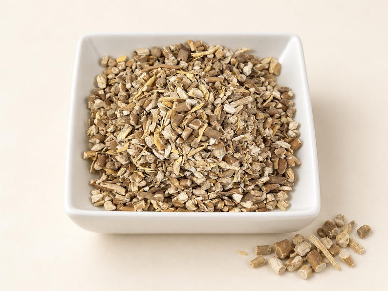 May include: A square white bowl filled with a mound of dried, chopped herbal root pieces. The pieces are a mix of light tan, beige, and dark brown woody textures. A small pile of the same dried root fragments sits outside the bowl to the lower right.