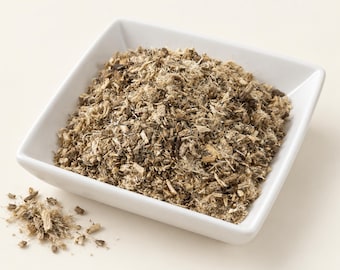 May include: A square white ceramic bowl filled with a pile of dried, shredded, light brown and dark brown fibrous plant bark. The texture is coarse and splintered, with small fragments scattered outside the bowl on the surface.