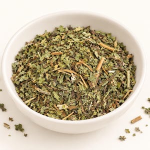 May include: A white ceramic bowl filled with a blend of dried green herbs, including small crinkled leaves and thin woody stems. The mixture features various shades of olive, sage, and dark green, with some scattered pieces resting outside the bowl.