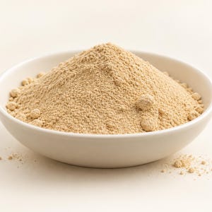 Könnte beinhalten: Ein Haufen feines, hellbeiges Pulver, das hoch in einer glatten, weißen Keramikschale aufgetürmt ist. Die Textur des Pulvers wirkt trocken und körnig, mit einigen kleinen Klumpen auf der Oberfläche und verstreuten Partikeln am Boden der Schale.