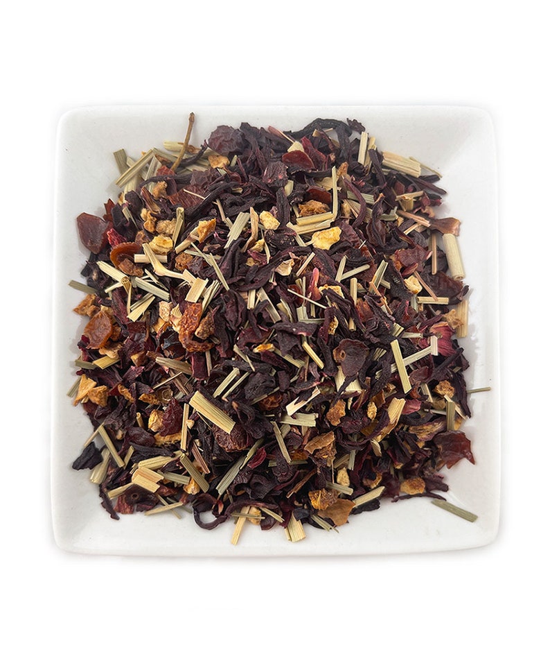 May include: A close-up of a white square dish filled with a blend of dried hibiscus flowers, orange peel, and lemongrass. The mixture is a deep red color with hints of orange and yellow.