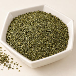 May include: A white hexagonal bowl filled with a mound of finely chopped, dried dark green herbs. The texture of the herb pieces is coarse and granular, with some small scattered particles visible outside the bowl on the surface.