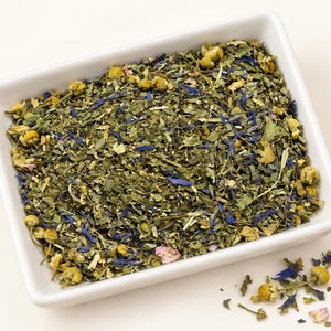 May include: A white rectangular bowl filled with a loose leaf herbal tea blend containing dried green leaves, small yellow chamomile flowers, vibrant blue cornflower petals, and tiny pink flower fragments, with a few pieces scattered outside.