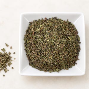 May include: A square white ceramic bowl filled with a mound of dried, crushed green herbs. The herbs are a mix of dark and light green shades with small, jagged leaf fragments. A small, loose pile of the same dried herbs sits to the left of the bowl.