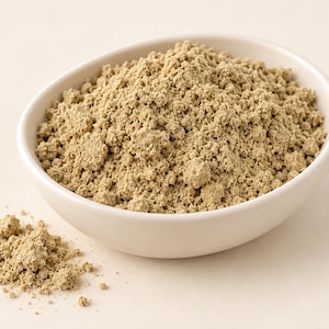 May include: A small, off-white ceramic bowl filled with a mound of fine, light-brown, earthy-toned powder. A small portion of the same textured, granular powder is scattered outside the bowl, showing a consistent, dry, and matte appearance.