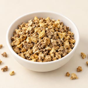 May include: A white ceramic bowl filled with small, irregular, dried tan and light brown root pieces. The texture of the pieces is rough and fibrous, with a mix of pale yellow and earthy brown tones throughout the pile, and a few scattered pieces.