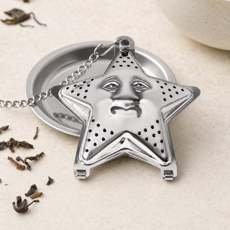 May include: A silver-colored stainless steel tea infuser shaped like a five-pointed star. The front surface features a molded face with eyes, a nose, and a mouth, and is perforated with small holes. A metal chain is attached to the top point.