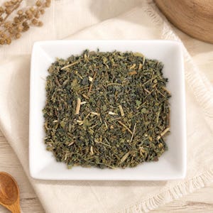 May include: A square white ceramic bowl filled with a mixture of dried green herbs, leaves, and small woody stems. The texture of the herbal blend is coarse and varied, featuring shades of dark forest green, olive, and light tan throughout the mix.