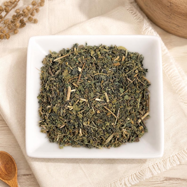 May include: A square white ceramic bowl filled with a mixture of dried green herbs, leaves, and small woody stems. The texture of the herbal blend is coarse and varied, featuring shades of dark forest green, olive, and light tan throughout the mix.