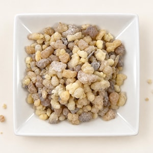May include: A square white bowl filled with a collection of small, irregular, translucent resin pieces in shades of pale yellow, cream, and dark brown. The resin chunks have a crystalline, textured appearance and are piled together in the center.