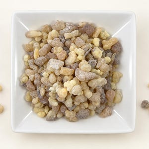 May include: A square white ceramic bowl filled with a collection of small, irregular, translucent resin pieces in shades of pale yellow, beige, and light brown. Several individual resin chunks are scattered outside the bowl on the surface.