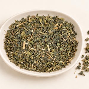 May include: A white ceramic bowl filled with a mixture of dried green herbs, including small dark green leaves and light tan woody stems. A small pile of the same dried herbal mixture is scattered to the right of the bowl on the surface.