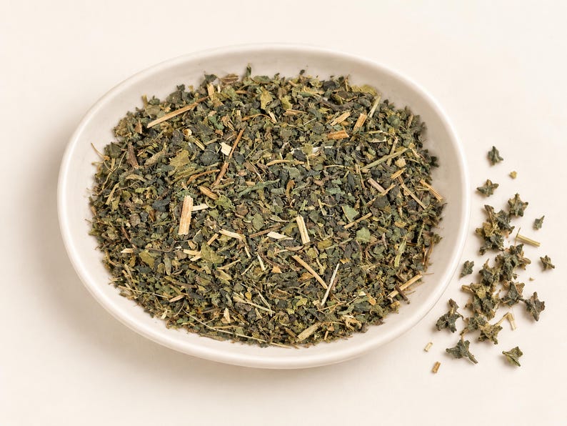 May include: A white ceramic bowl filled with a mixture of dried green herbs, including small dark green leaves and light tan woody stems. A small pile of the same dried herbal mixture is scattered to the right of the bowl on the surface.