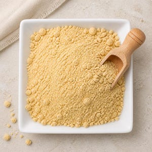 May include: A square white ceramic bowl filled with fine, pale yellow powder. A small, light-coloured wooden scoop rests inside the bowl, partially submerged in the powder. The texture of the powder appears slightly granular with small clumps.