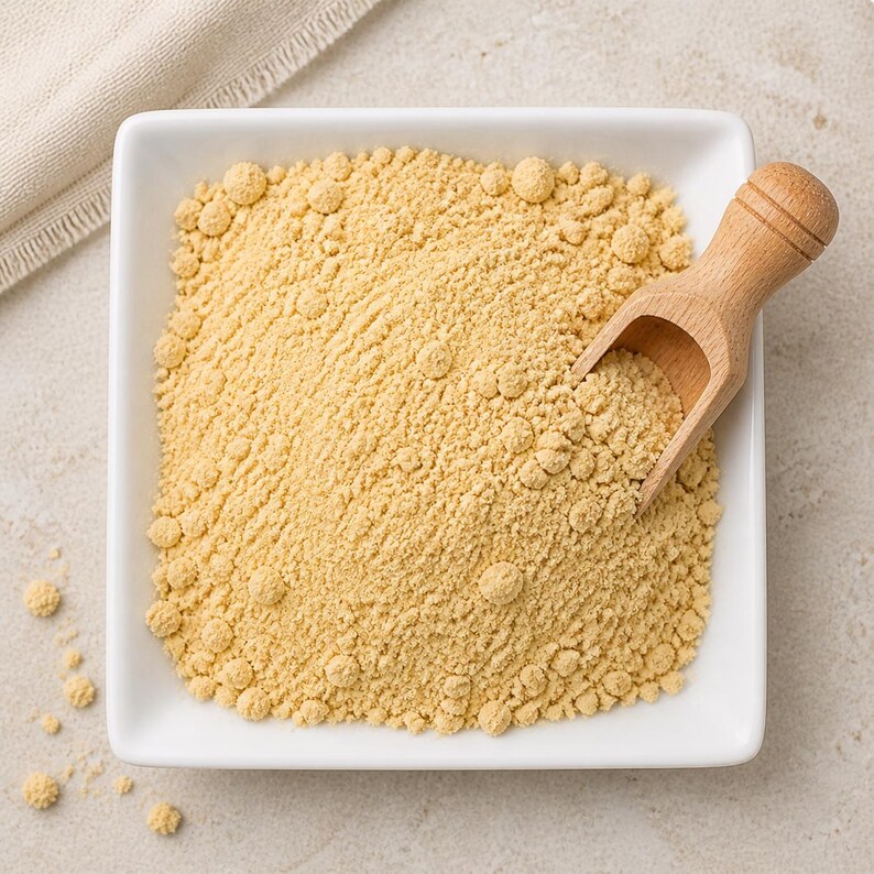 May include: A square white ceramic bowl filled with fine, pale yellow powder. A small, light-coloured wooden scoop rests inside the bowl, partially submerged in the powder. The texture of the powder appears slightly granular with small clumps.