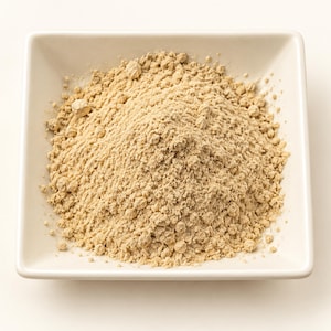 May include: A mound of fine, light beige powder is centered in a square, white ceramic bowl. The powder has a granular, textured appearance with small clumps scattered throughout the pile, contrasting against the smooth, solid white surface.