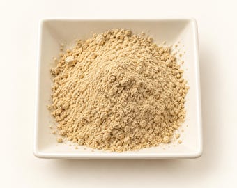 May include: A mound of fine, light beige powder is centered in a square, white ceramic bowl. The powder has a granular, textured appearance with small clumps scattered throughout the pile, contrasting against the smooth, solid white surface.