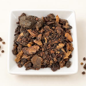 May include: A square white bowl filled with a collection of dark brown, resinous, and woody chunks. The pieces vary in size and texture, displaying shades of deep brown, tan, and black. Several loose fragments are scattered outside the bowl.