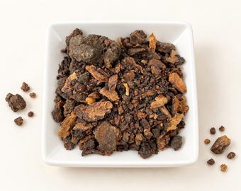 May include: A square white bowl filled with a collection of dark brown, resinous, and woody chunks. The pieces vary in size and texture, displaying shades of deep brown, tan, and black. Several loose fragments are scattered outside the bowl.