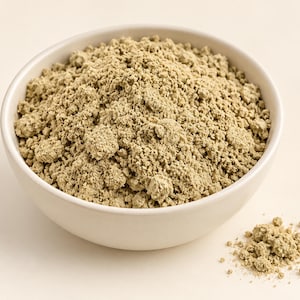 May include: A round, off-white ceramic bowl filled with a mound of fine, light olive-green powder. The texture of the powder is slightly clumpy and granular. A small pile of the same light olive-green powder sits outside the bowl to the right.