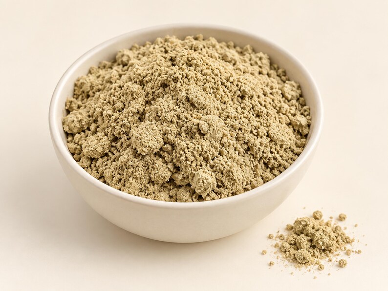 May include: A round, off-white ceramic bowl filled with a mound of fine, light olive-green powder. The texture of the powder is slightly clumpy and granular. A small pile of the same light olive-green powder sits outside the bowl to the right.