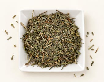 May include: A square white bowl filled with a blend of dried green herbs, including thin stems and small, fragmented leaves in various shades of olive, sage, and dark green. Several loose pieces of the dried herbal mixture are scattered outside.
