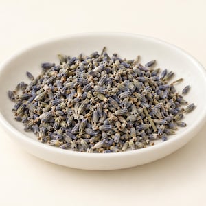 Puede incluir: Un pequeño plato redondo de cerámica blanca lleno de un montón de capullos de lavanda secos. La lavanda consiste en numerosos pétalos diminutos y alargados de color azul violáceo y pequeños tallos verdes, creando un montón orgánico y texturizado.