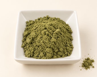 May include: A square white ceramic bowl filled with a mound of fine, olive-green powder. The texture of the powder appears dry and slightly clumped, with a small amount of the same green substance scattered outside the bowl on the surface.