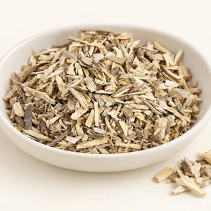 May include: A white ceramic bowl filled with a mound of dried, shredded light brown and tan wood bark pieces. A small pile of the same shredded bark fragments is scattered on the surface next to the bowl, showing fibrous textures and earthy tones.