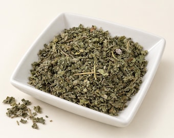 May include: A square white ceramic bowl filled with dried green herbal leaves and small stems. The texture of the dried herbs is coarse and fragmented, showing various shades of olive and dark green. A small pile of the same dried herbs sits nearby.