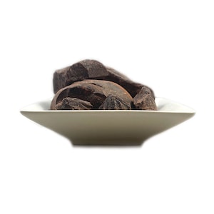 May include: A small, white, square-shaped bowl filled with several irregular, dark brown chunks of solid chocolate. The pieces have a rough, matte texture and are piled together in the center of the shallow dish, creating a contrast in color.