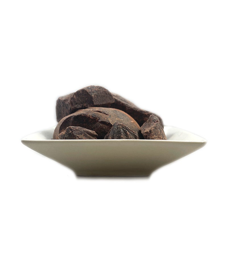 May include: A small, white, square-shaped bowl filled with several irregular, dark brown chunks of solid chocolate. The pieces have a rough, matte texture and are piled together in the center of the shallow dish, creating a contrast in color.