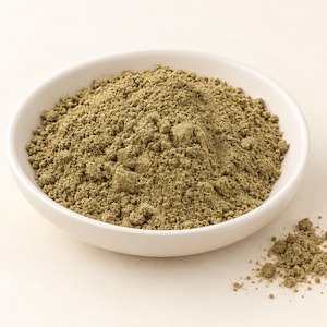 May include: A white ceramic bowl filled with a mound of fine, earthy, olive-green powder. The texture of the powder is granular and dry, with a small amount of the same green substance scattered outside the bowl on the surface.