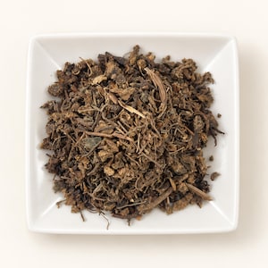 May include: A square white ceramic dish filled with a mound of dried, dark brown herbal tea leaves. The mixture consists of small, fragmented pieces of roots, stems, and dark foliage, creating a textured, earthy appearance in the center of the bowl.