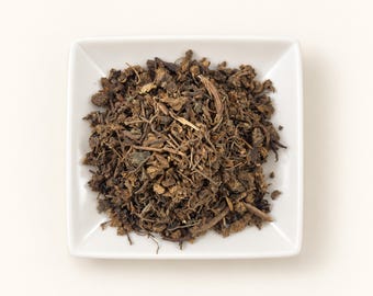 May include: A square white ceramic dish filled with a mound of dried, dark brown herbal tea leaves. The mixture consists of small, fragmented pieces of roots, stems, and dark foliage, creating a textured, earthy appearance in the center of the bowl.