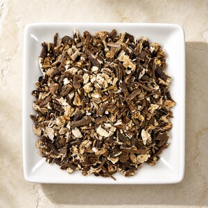 May include: A square white ceramic bowl filled with a heap of dried, chopped brown and tan root pieces. The pieces vary in size and texture, showing a mix of dark brown bark-like exteriors and lighter, fibrous interiors throughout the pile.
