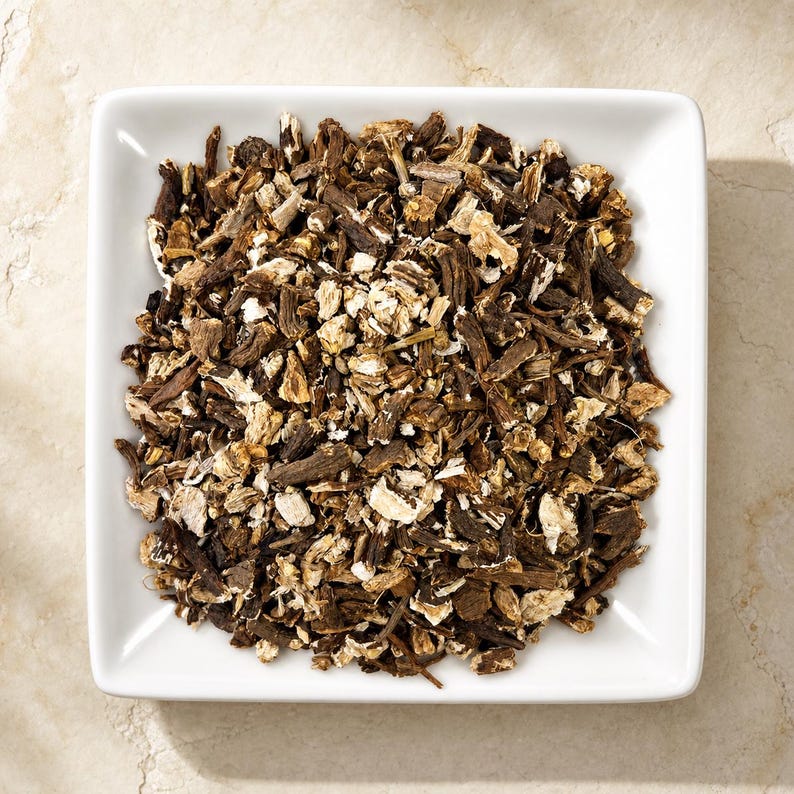 May include: A square white ceramic bowl filled with a heap of dried, chopped brown and tan root pieces. The pieces vary in size and texture, showing a mix of dark brown bark-like exteriors and lighter, fibrous interiors throughout the pile.