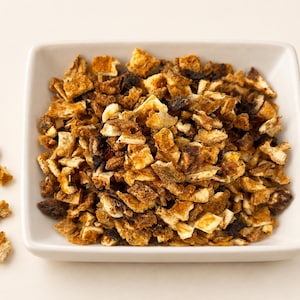 May include: A square white bowl filled with small, dried orange peel pieces. The pieces are irregular in shape and feature a textured surface with shades of golden yellow, burnt orange, and dark brown. Several loose pieces are scattered nearby.