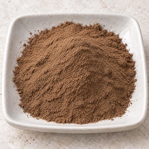 May include: A mound of fine, medium-brown powder contained within a square, off-white ceramic dish with rounded corners. The texture of the powder appears uniform and dry, forming a conical shape that fills the center of the shallow bowl.