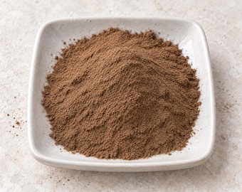May include: A mound of fine, medium-brown powder contained within a square, off-white ceramic dish with rounded corners. The texture of the powder appears uniform and dry, forming a conical shape that fills the center of the shallow bowl.