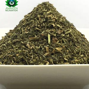 May include: A close-up of a white bowl filled with a large pile of dried green herbs. The herbs are a mix of different sizes and shapes, and they appear to be finely chopped.