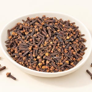 May include: A white ceramic bowl filled with a large quantity of dried whole cloves. The cloves are dark brown with lighter tan, rounded flower buds at the tips. Several individual dried cloves are scattered outside the bowl on the surface.