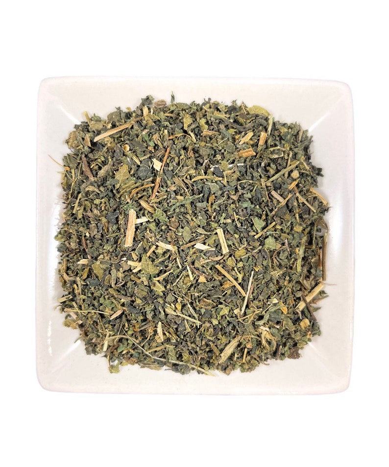 May include: A square white ceramic dish filled with a pile of dried, crushed green herbal leaves and small woody stems. The mixture consists of various shades of dark and olive green foliage, interspersed with light tan, fibrous plant fragments.