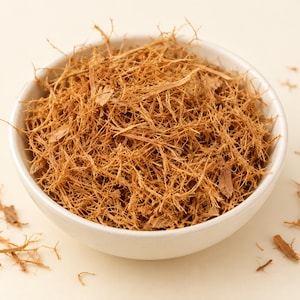 May include: A white ceramic bowl filled with a mound of dried, shredded light brown bark fibers. The texture of the material is thin, woody, and tangled, with several small loose pieces scattered around the base of the bowl on a light surface.