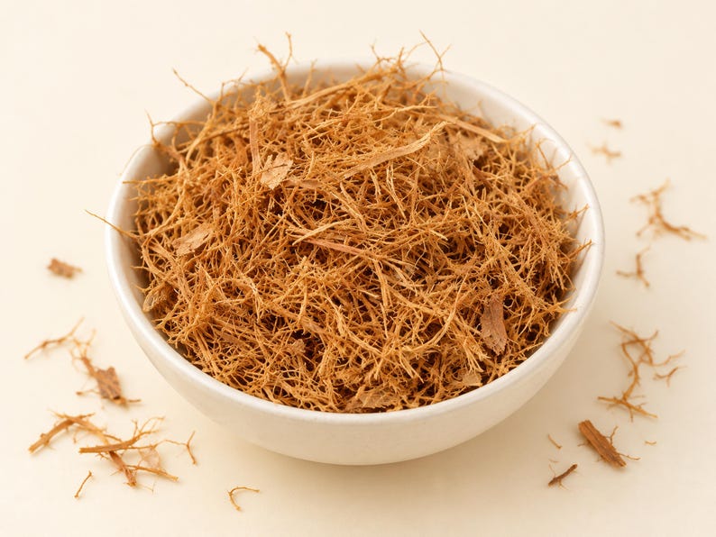 May include: A white ceramic bowl filled with a mound of dried, shredded light brown bark fibers. The texture of the material is thin, woody, and tangled, with several small loose pieces scattered around the base of the bowl on a light surface.