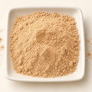 May include: A square white ceramic dish filled with a mound of fine, light tan powder. The powder has a dry, granular texture and is piled high in the center of the dish, with a small amount of loose particles scattered around the edges.