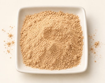 May include: A square white ceramic dish filled with a mound of fine, light tan powder. The powder has a dry, granular texture and is piled high in the centre of the dish, with a small amount of loose particles scattered around the edges.